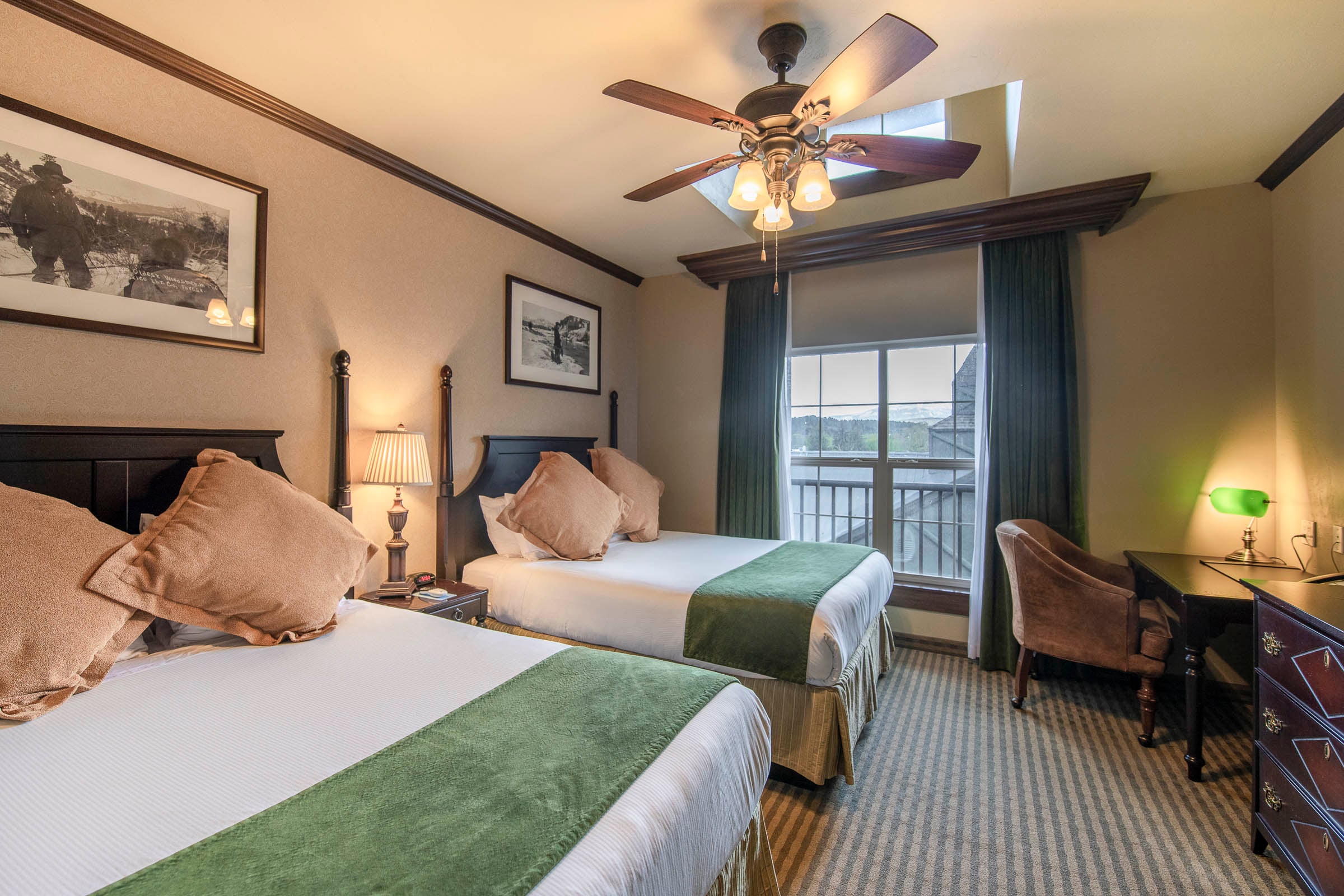 Two beds with white linens and green runners are centered in a hotel room. A ceiling fan is overhead, a window shows a balcony, and a desk with a green lamp is right. Text on framed artwork: Oldest homestead in the C___ break.