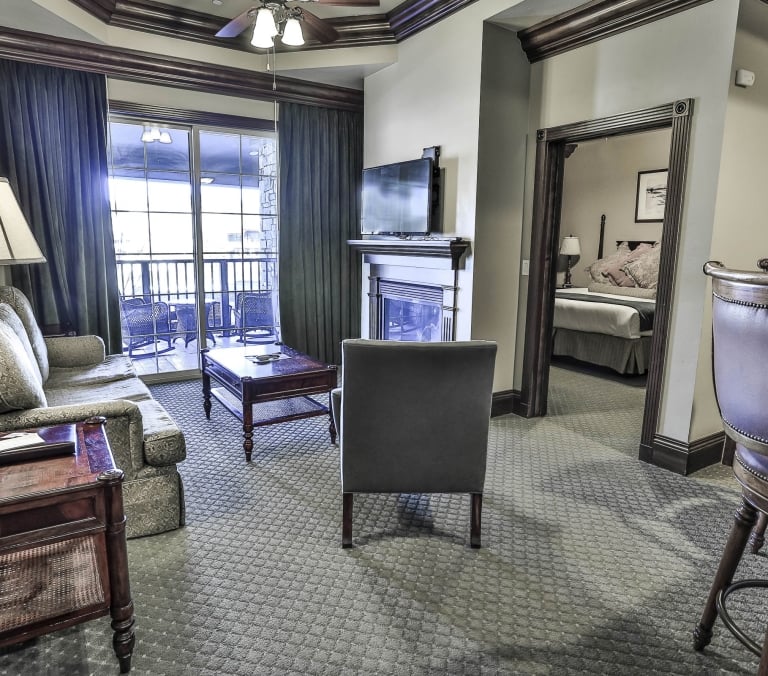 A hotel living room furnished with a sofa, coffee table, fireplace, and TV, opening to a balcony and a bedroom, featuring elegant decor. Text: 