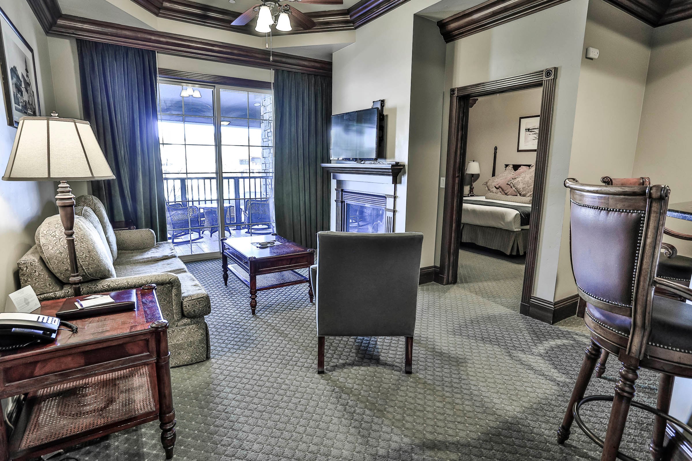 A hotel living room furnished with a sofa, coffee table, fireplace, and TV, opening to a balcony and a bedroom, featuring elegant decor. Text: 