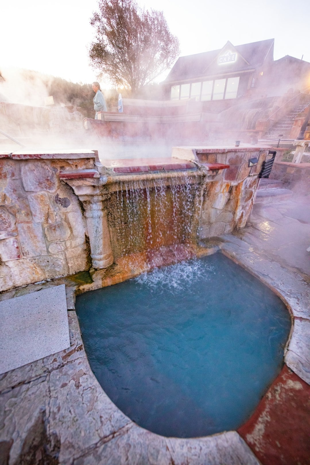 Water cascades from a stone wall into a blue hot spring pool, while steam rises from multiple pools. A person stands near an upper pool, with a building labeled CAFE in the background.