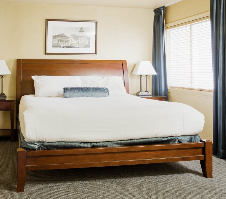 A queen-sized bed, neatly made with white linens and a blue bolster pillow, is flanked by nightstands with lamps in a hotel room with light yellow walls and a window.