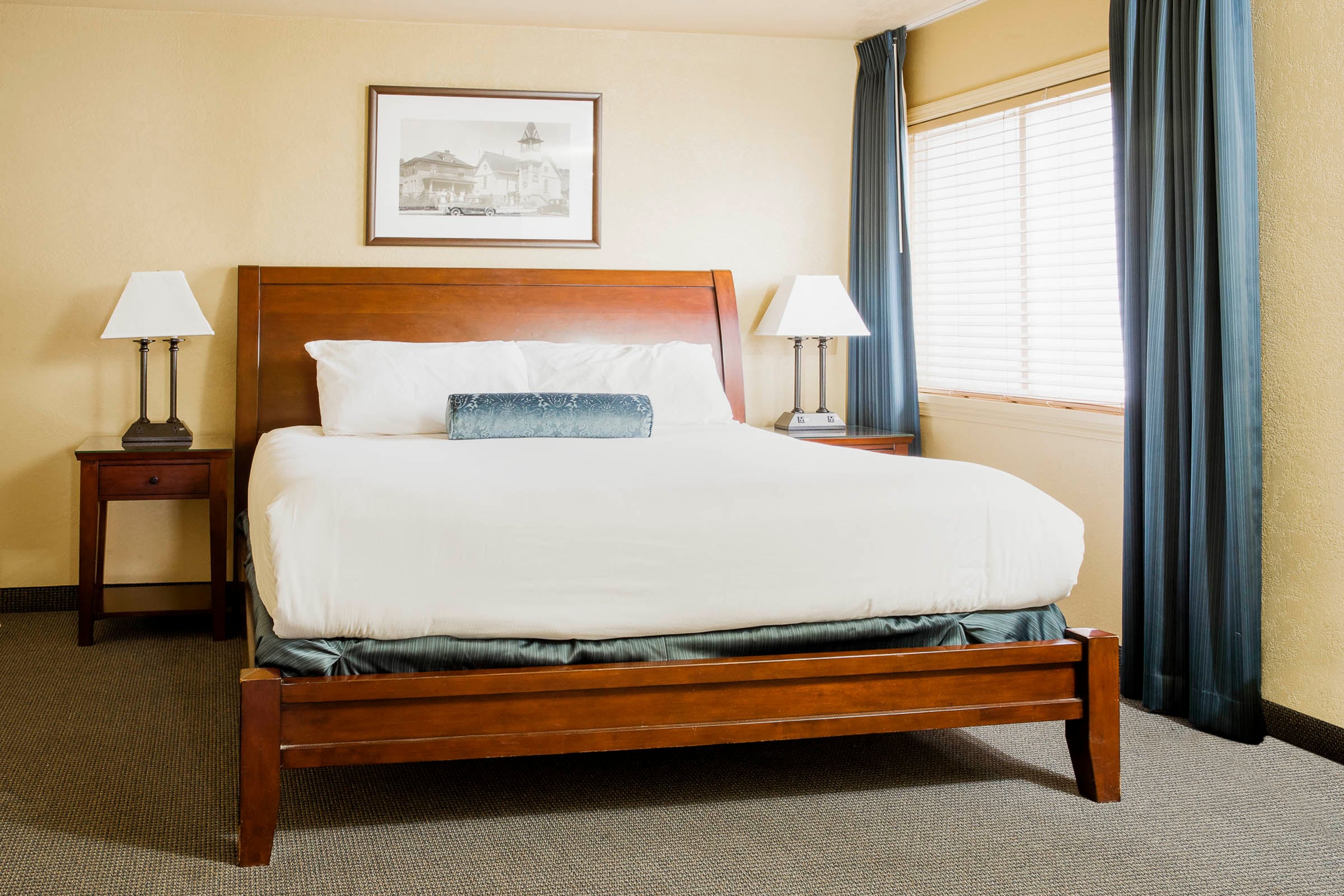 A queen-sized bed, neatly made with white linens and a blue bolster pillow, is flanked by nightstands with lamps in a hotel room with light yellow walls and a window.