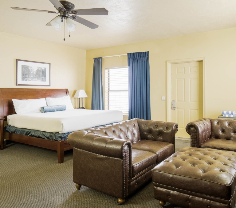 King-size bed and brown leather tufted sofa, armchair, and ottoman furnishing a hotel room with yellow walls, a ceiling fan, blue curtains, and patterned carpet.