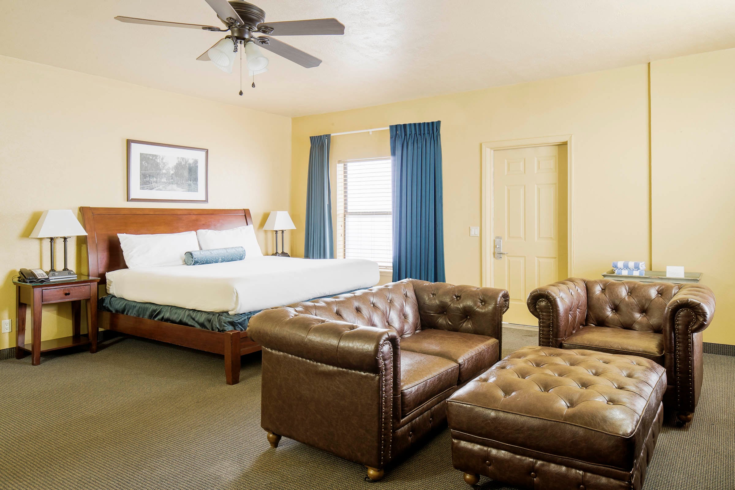 King-size bed and brown leather tufted sofa, armchair, and ottoman furnishing a hotel room with yellow walls, a ceiling fan, blue curtains, and patterned carpet.