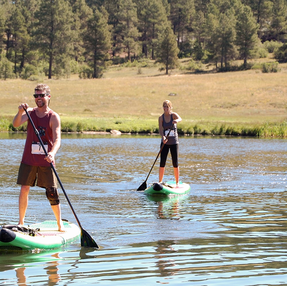 A man and woman stand-up paddleboard on a calm lake, surrounded by a grassy bank and a pine forest. The man wears bib 