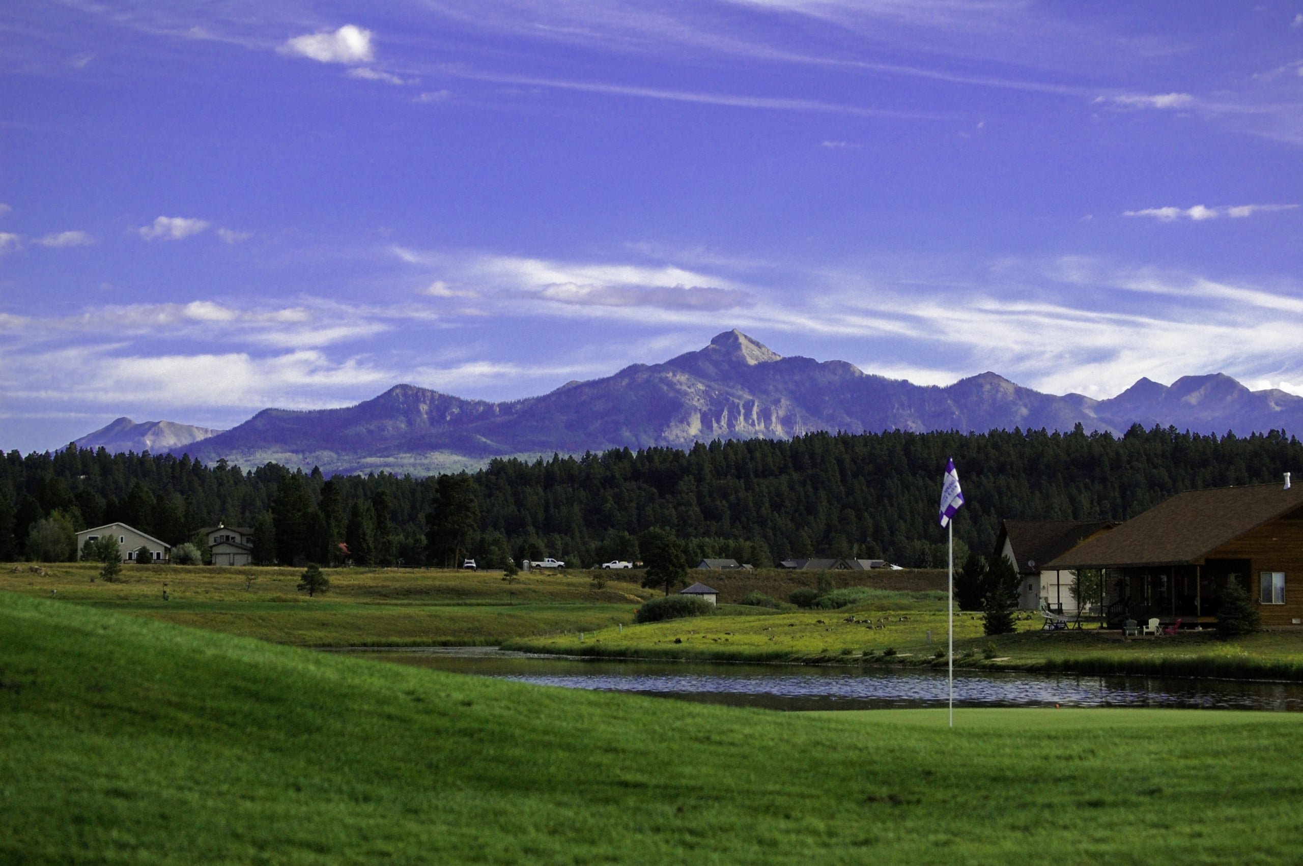A golf course green, pond, and houses are backed by a dense forest and majestic mountains under a blue sky. A purple and white flag, reading 