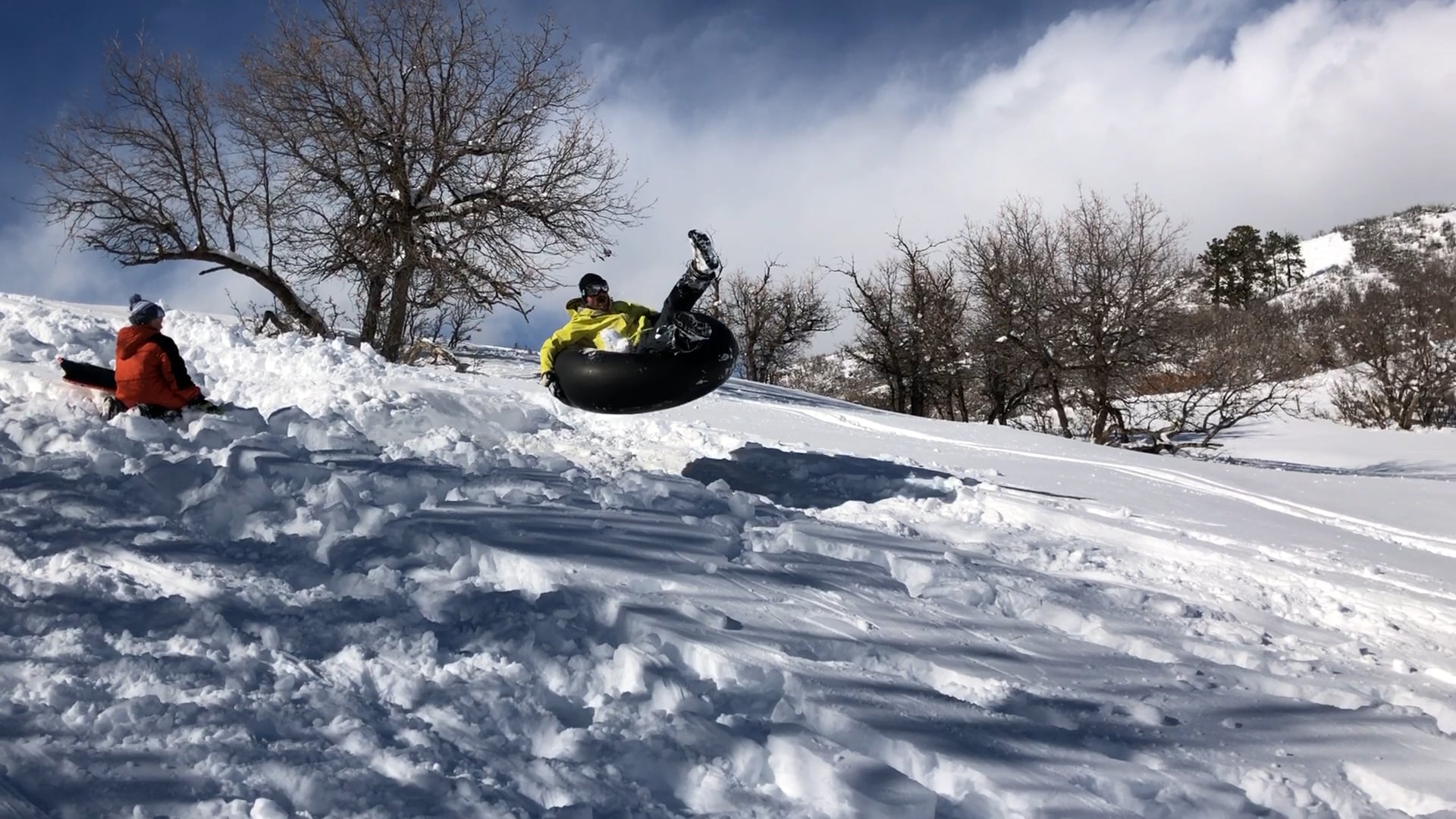A person in a yellow jacket is airborne on a black snow tube over a jump, with another person sitting on a sled in the snow-covered, treed landscape.