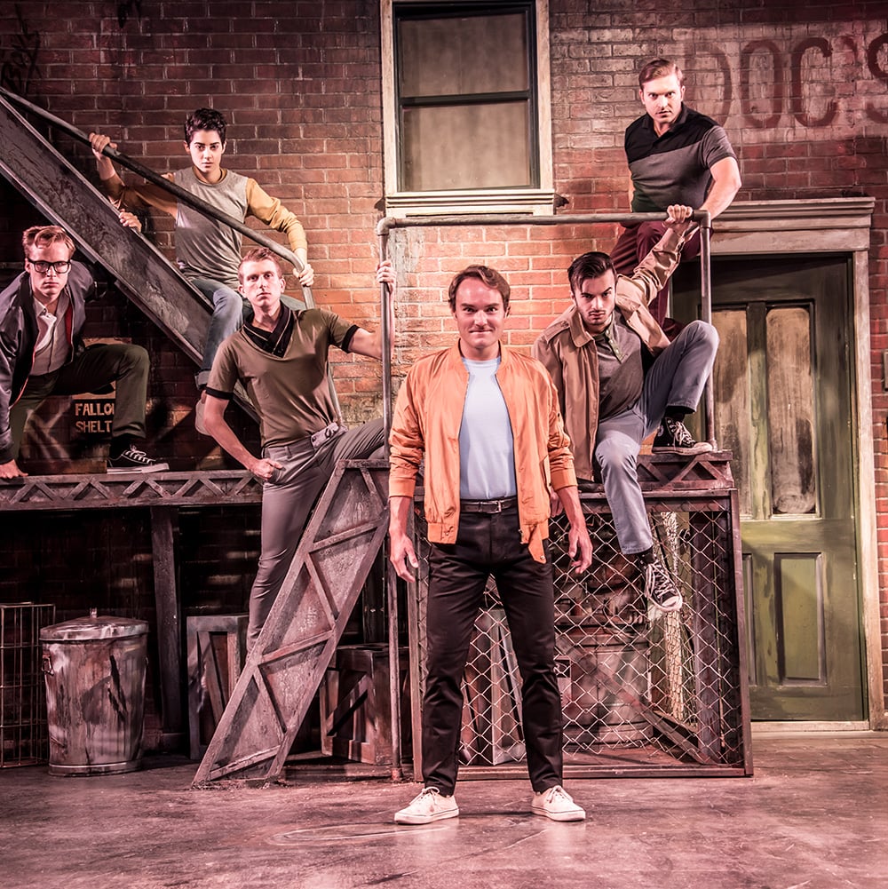 Six men pose on a brick alleyway set with a fire escape and platforms. One man stands center, others are seated or leaning. Text: DOC'S, FALLO, SHELT.