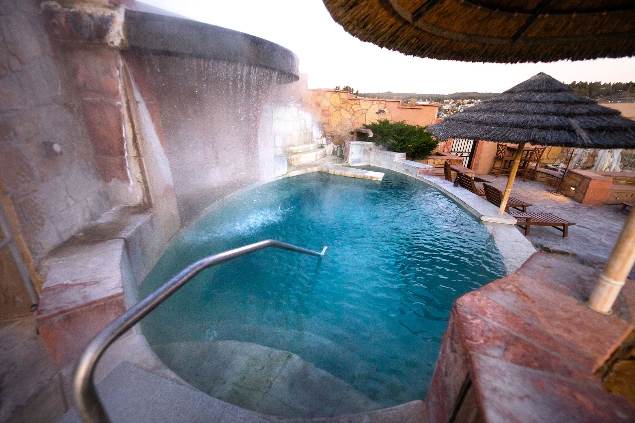 A steaming turquoise hot tub with cascading water from a stone waterfall, surrounded by stone walls and a thatched umbrella under an overcast sky.