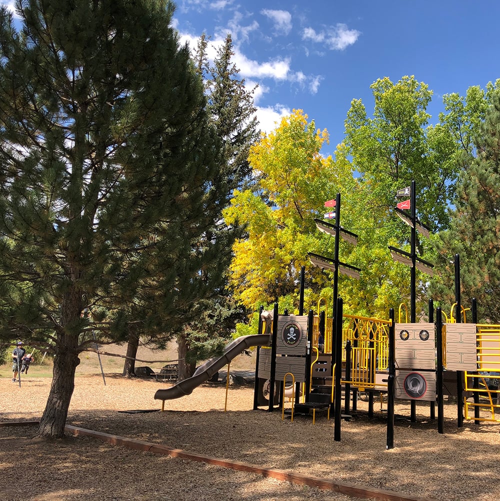 A pirate ship playground structure offers slides and climbing areas. It is surrounded by tall trees (green and yellow) on wood chips under a blue sky. Text: PIRATE, skull and crossbones.