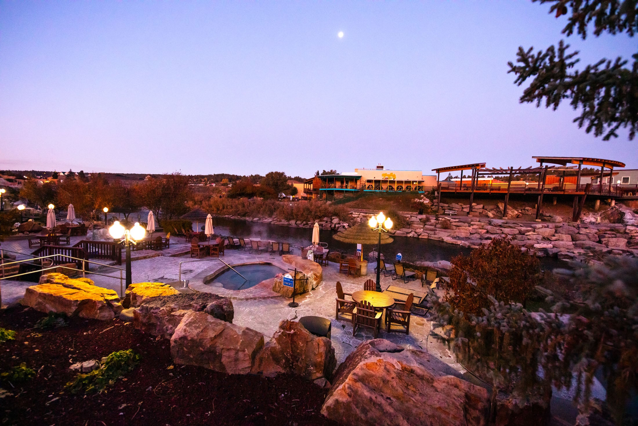 Outdoor hot springs resort at twilight, featuring multiple illuminated pools and seating areas. Buildings are visible under a moonlit purple sky. Text: PLEASE, ENTER HERE.