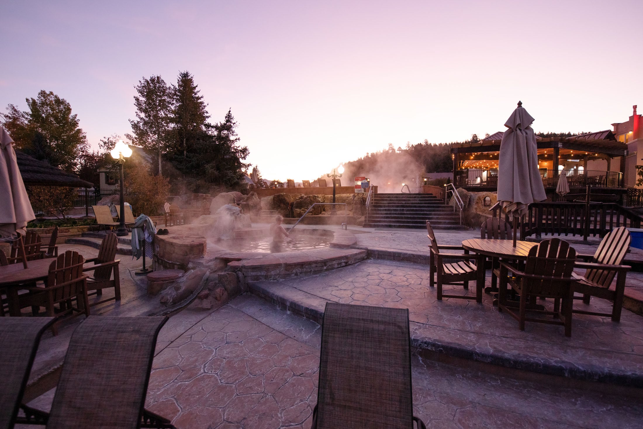 Steaming hot springs complex where people soak in multiple pools. Surrounded by lounge areas, resort buildings, and trees under a purple twilight sky. Text: Water Quality Posting.