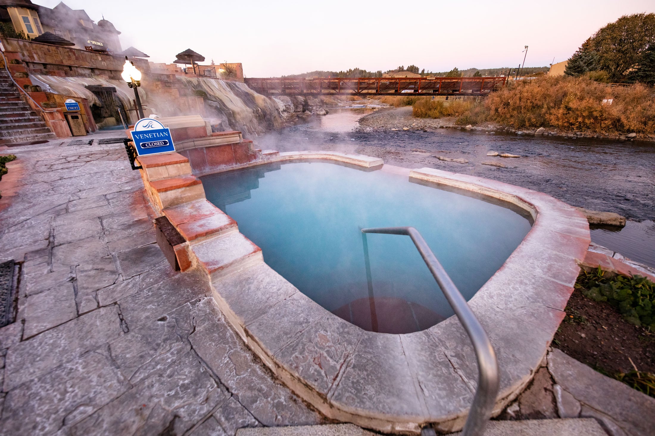 A steaming, turquoise hot springs pool sits beside a river with a pedestrian bridge, surrounded by stone paving and a tiered building. Signs read: VENETIAN CLOSED, 4 SPRINGS, FRESH TOWELS.
