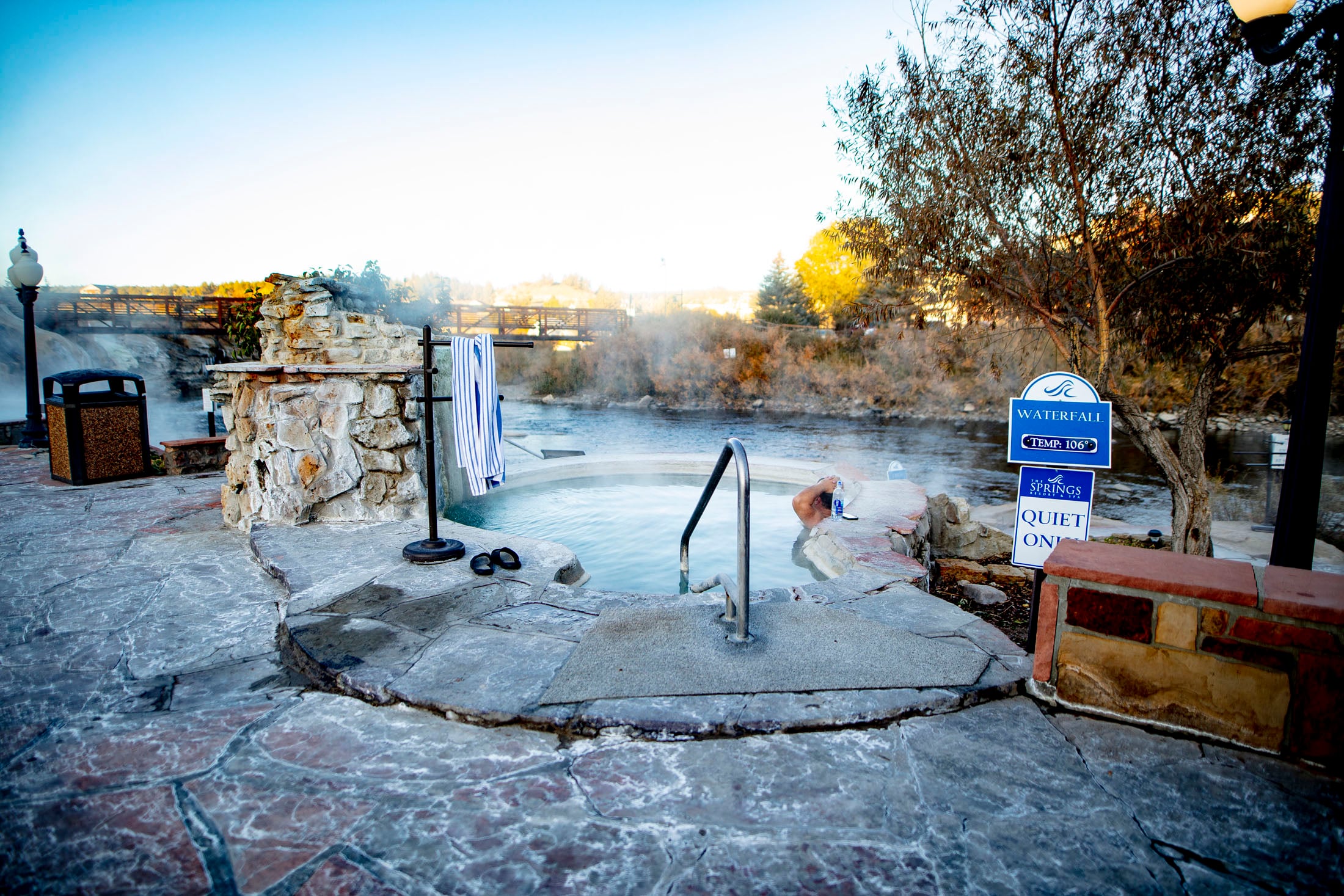 A steaming outdoor hot tub with a person relaxing, set in a stone patio beside a river under a clear sky, with a bridge in the distance. Text: WATERFALL TEMP: 106°, THE SPRINGS RESORT & SPA, QUIET ONE.