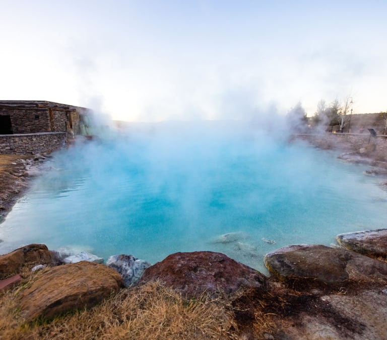 A vibrant blue hot spring pool heavily steaming, surrounded by dry grass, large rocks, and a stone wall leading to a building with a wooden pergola under a pale sky.