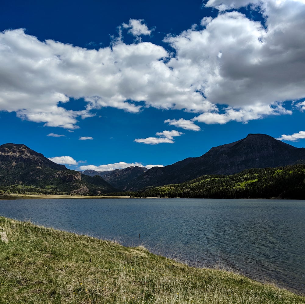A blue lake sits amidst dark, tree-covered mountains under a bright blue sky with scattered white clouds. Dry grass fills the foreground.