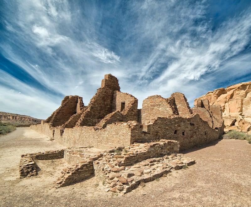 Vast, partially collapsed stone ruins, with visible stacked walls, stretch across an arid valley. A dirt path and rocky mesa flank the site under a blue sky with wispy clouds.