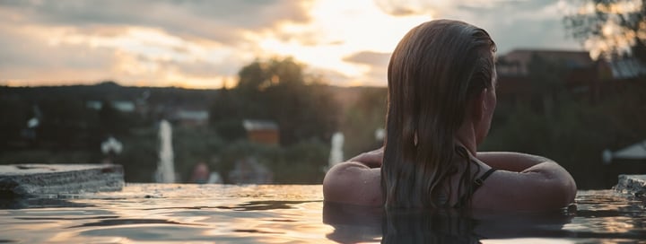 A woman with wet hair looks out from an infinity pool over a distant landscape at sunset, with warm light reflecting on the water.