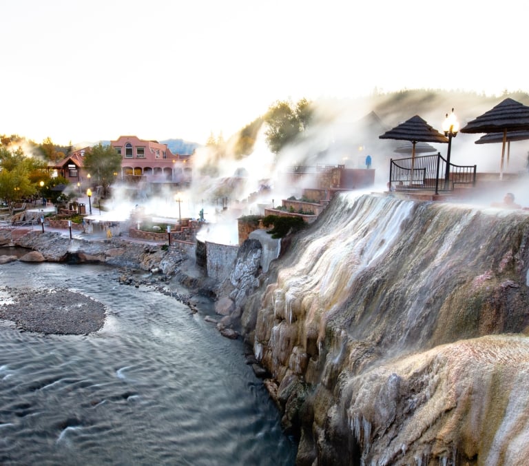 People bathe in steaming outdoor hot springs, cascading over mineral-stained cliffs beside a flowing river. Resort buildings and trees line the banks under a clear sky.
