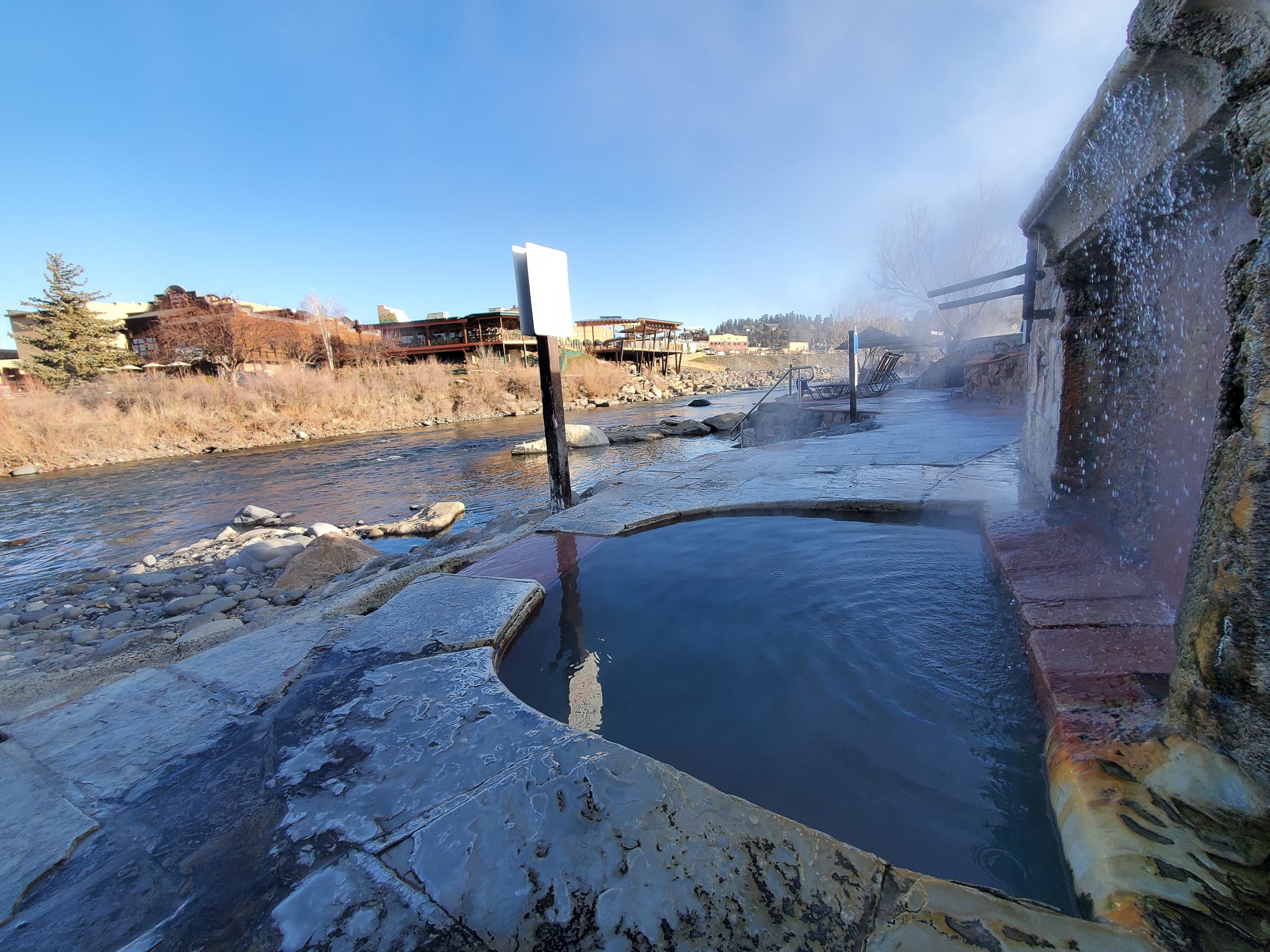 A steaming, curved hot spring pool receives cascading water from a rock wall, situated by a river with distant buildings and trees under a clear blue sky.