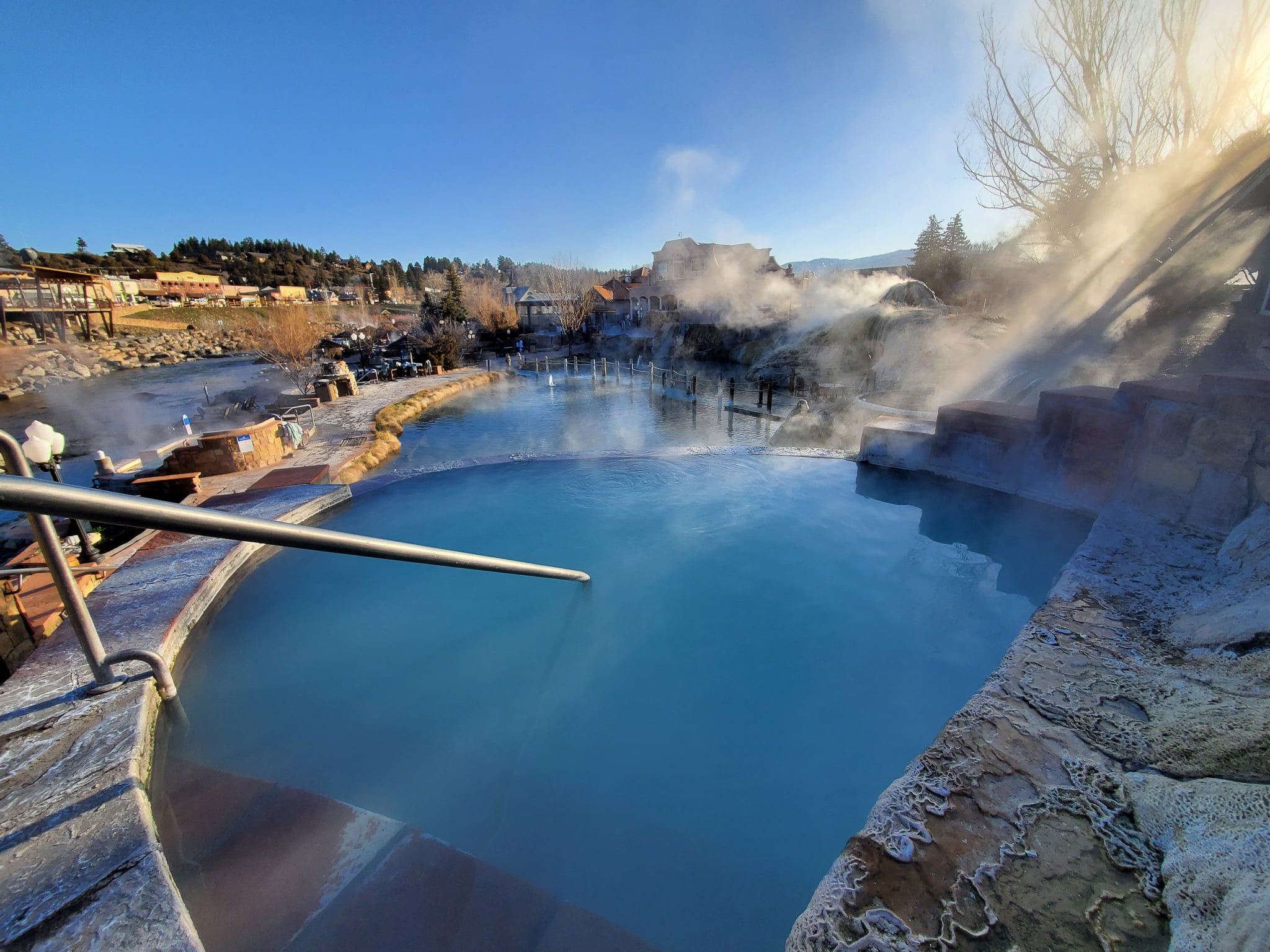 A large turquoise hot spring pool emits steam, sitting by a river with buildings and bare trees on a distant hillside under a clear blue sky.