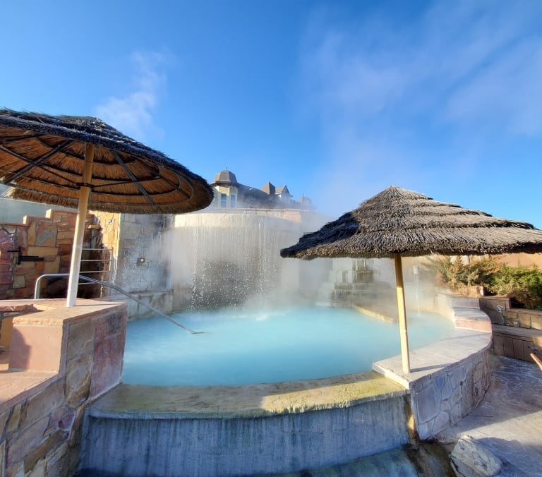 Steaming light blue hot spring pool with water cascading into it, flanked by two thatched umbrellas, under a clear blue sky with a building in the background.