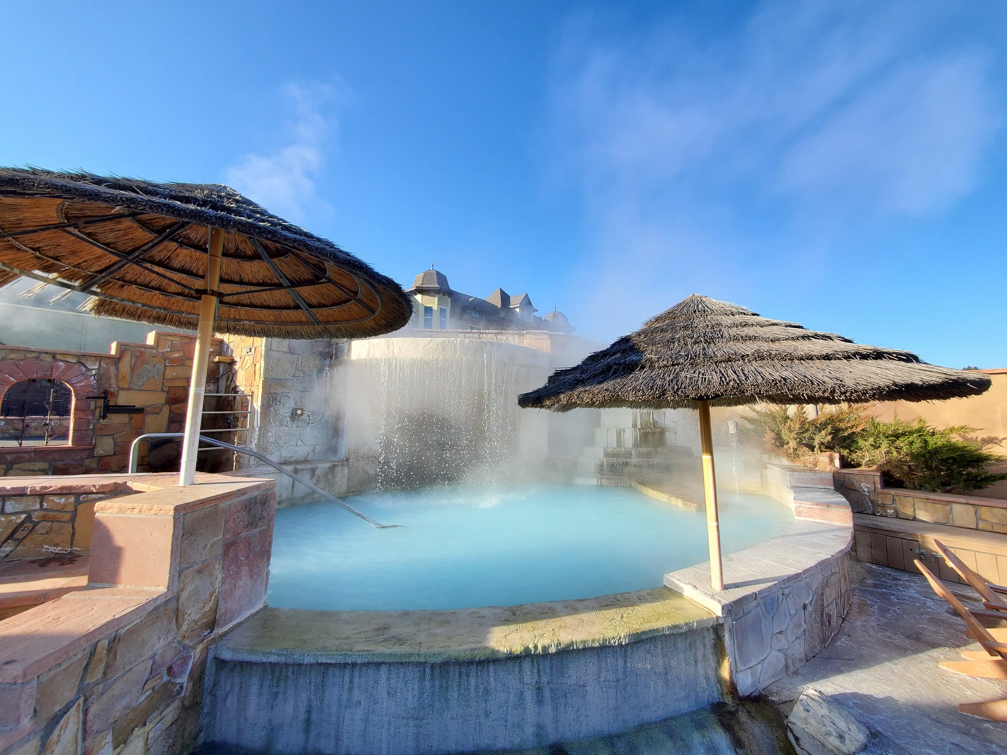 Steaming light blue hot spring pool with water cascading into it, flanked by two thatched umbrellas, under a clear blue sky with a building in the background.