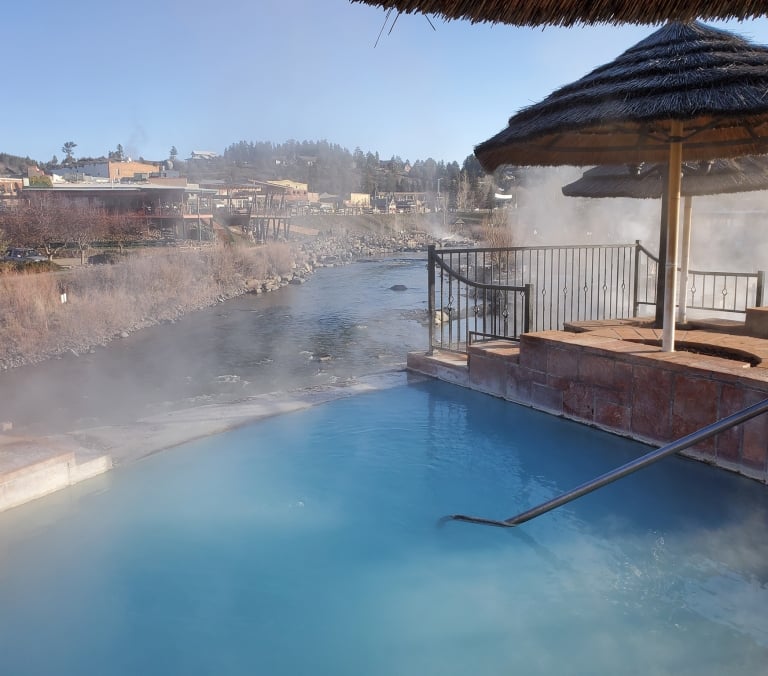A blue-hued hot spring pool is steaming, with a metal handrail. It lies next to a river, with a town containing a green 