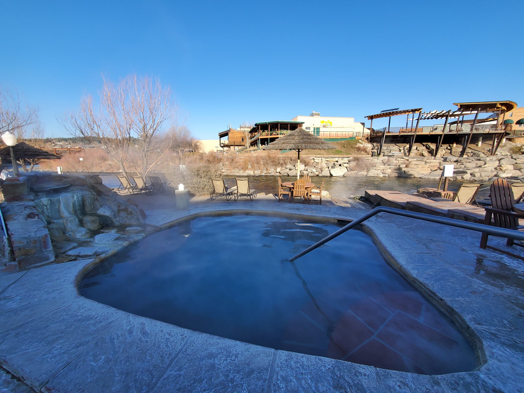 A steaming, heart-shaped hot spring pool with a handrail is set on a stone patio. A river, bare trees, and buildings are visible under a clear blue sky.