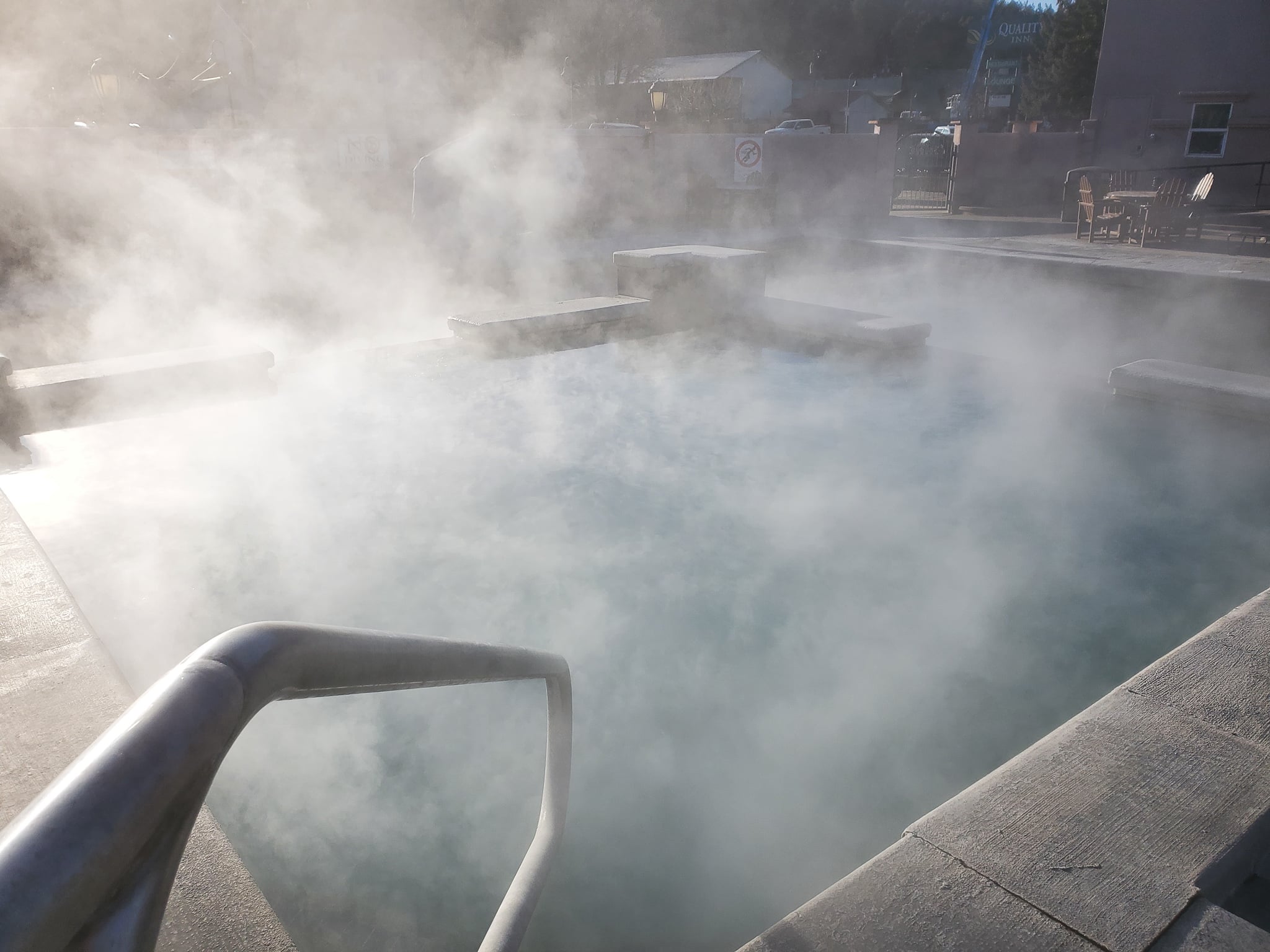 Outdoor pool intensely steaming, featuring submerged benches and a foreground handrail. Buildings in the background show a 