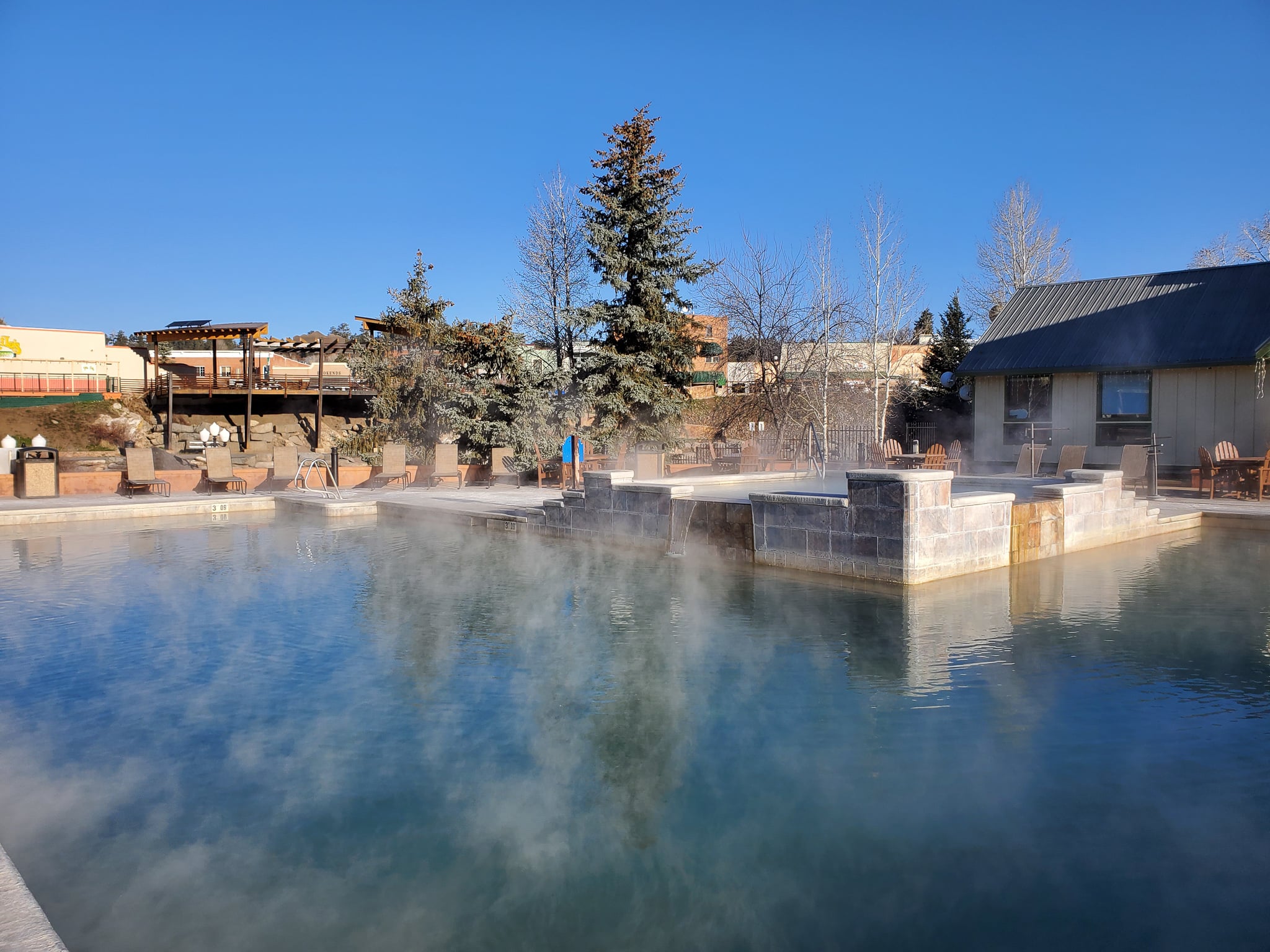 A large outdoor pool emits steam from its blue water, bordered by lounge chairs, trees, and buildings under a clear sky. Text reads: 