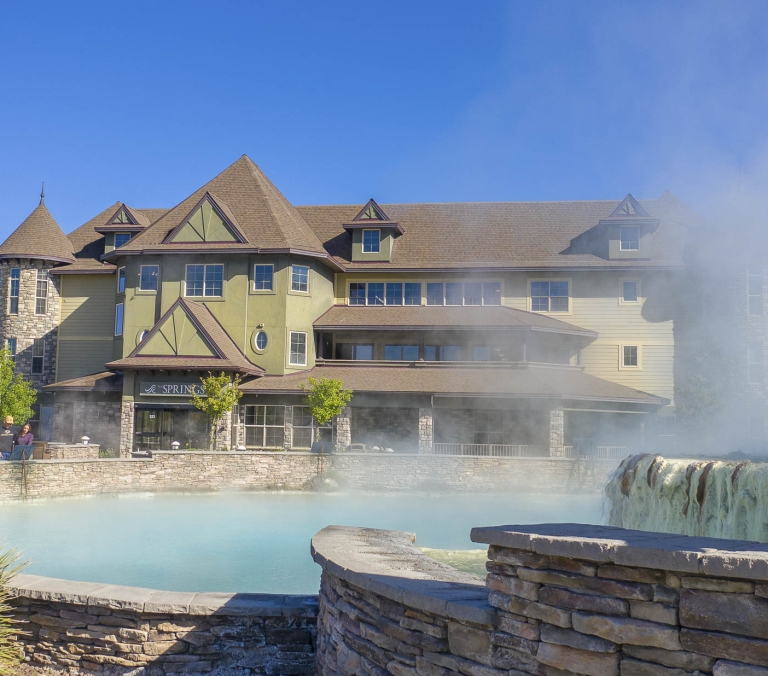 A lodge-style building overlooks a steaming blue mineral pool featuring a mineral-coated waterfall. People are visible near the pool under a clear blue sky. The SPRINGS 523.