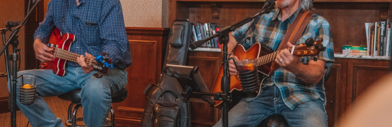 Two older men, one with a cowboy hat, are seated, playing guitars and singing into microphones. They perform indoors before a bookshelf. Text: THE HILL TO PAY, THINKING, FAST AND SLOW, RED BOOK, THIS IS HOW WE DO IT, A GIRL IN THE WOODS, THE NO. 1 LADIES DETECTIVE AGENCY, LAST WILL, TO KILL A MOCKINGBIRD, CALIFORNIA.