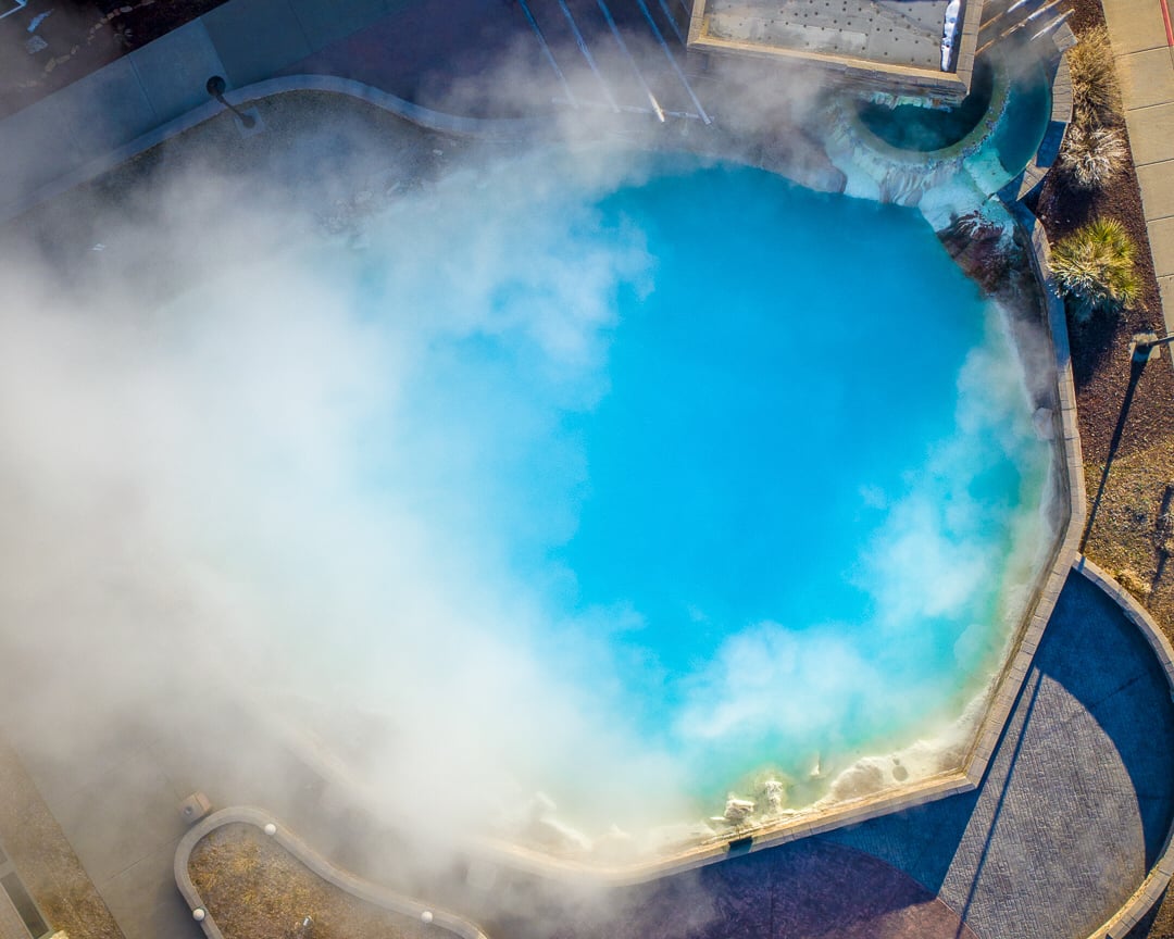 Large, bright blue geothermal pool steaming profusely, bordered by paved walkways and dry landscaping, viewed from above.