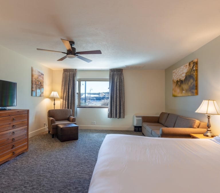 A hotel bedroom features a large bed, sofa, dresser with TV, and armchair. A ceiling fan hangs above, with a window revealing a snowy outdoor landscape.
