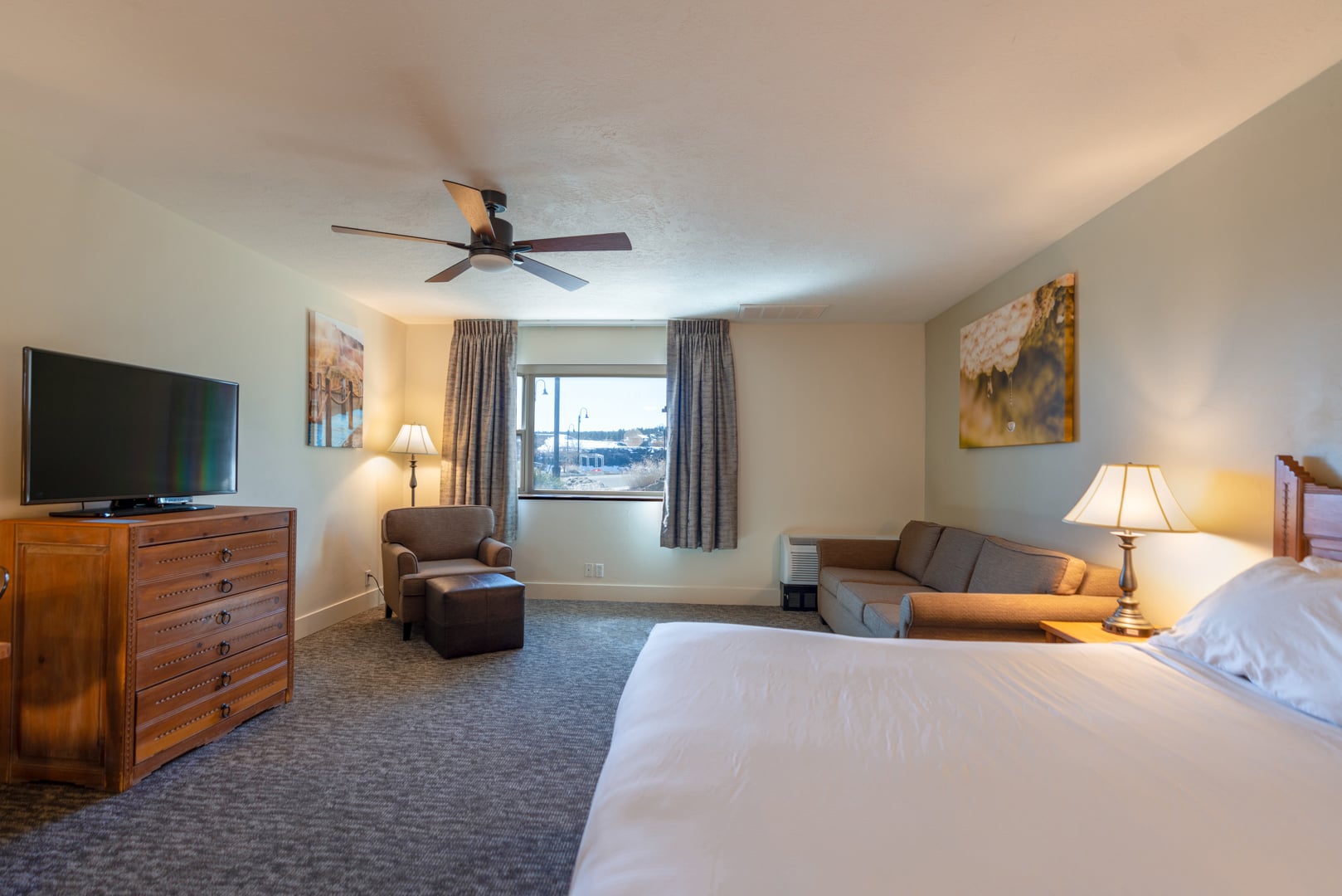 A hotel bedroom features a large bed, sofa, dresser with TV, and armchair. A ceiling fan hangs above, with a window revealing a snowy outdoor landscape.