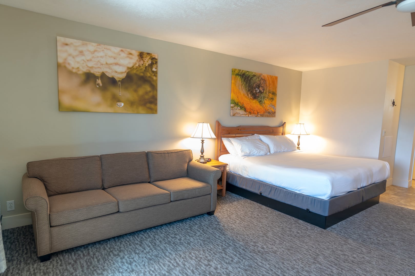 A brown sofa and a neatly made bed with white linens are illuminated by bedside lamps. Abstract art adorns the pale walls of this carpeted room.