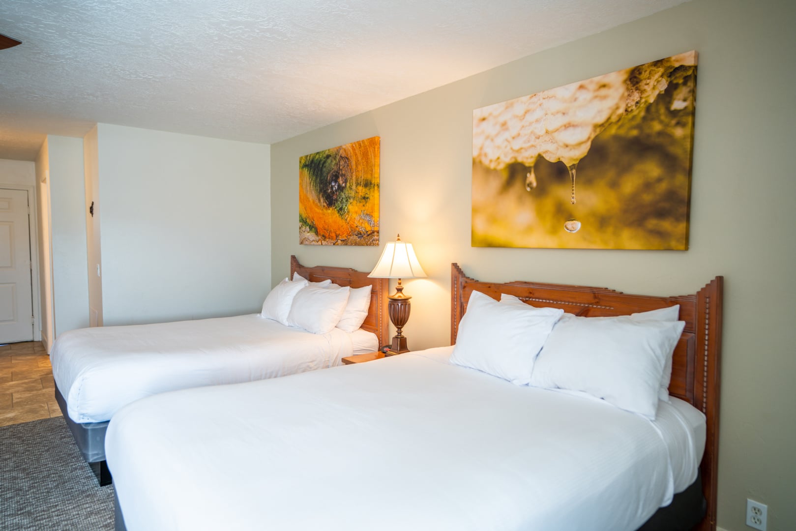 Two queen-sized beds with white linens and wooden headboards fill a hotel room, featuring a bedside lamp and two large abstract artworks on light green walls.