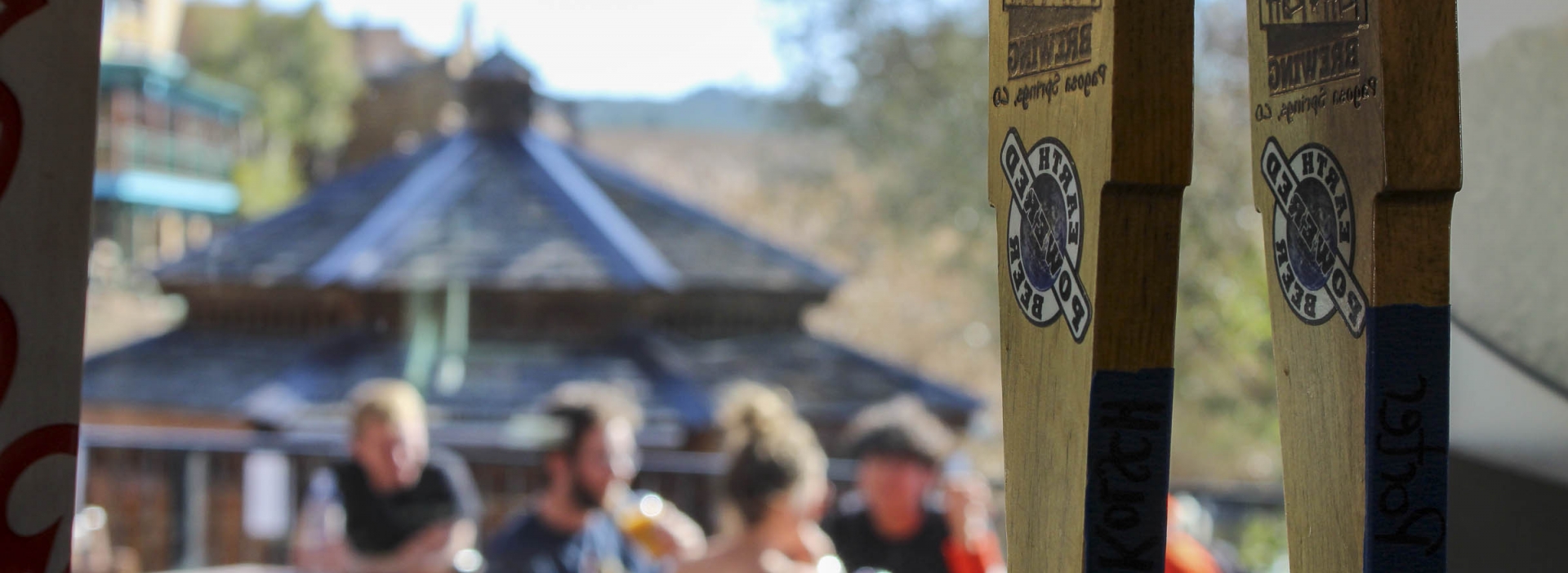 A close up of the hanging wooden decor of the Riff Raff Brewing overlooking a blurred image of a group drinking outdoors