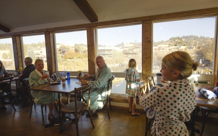 Several people in bathrobes are seated at wooden tables, some eating, while children stand near large windows, overlooking a sunny town and a river.