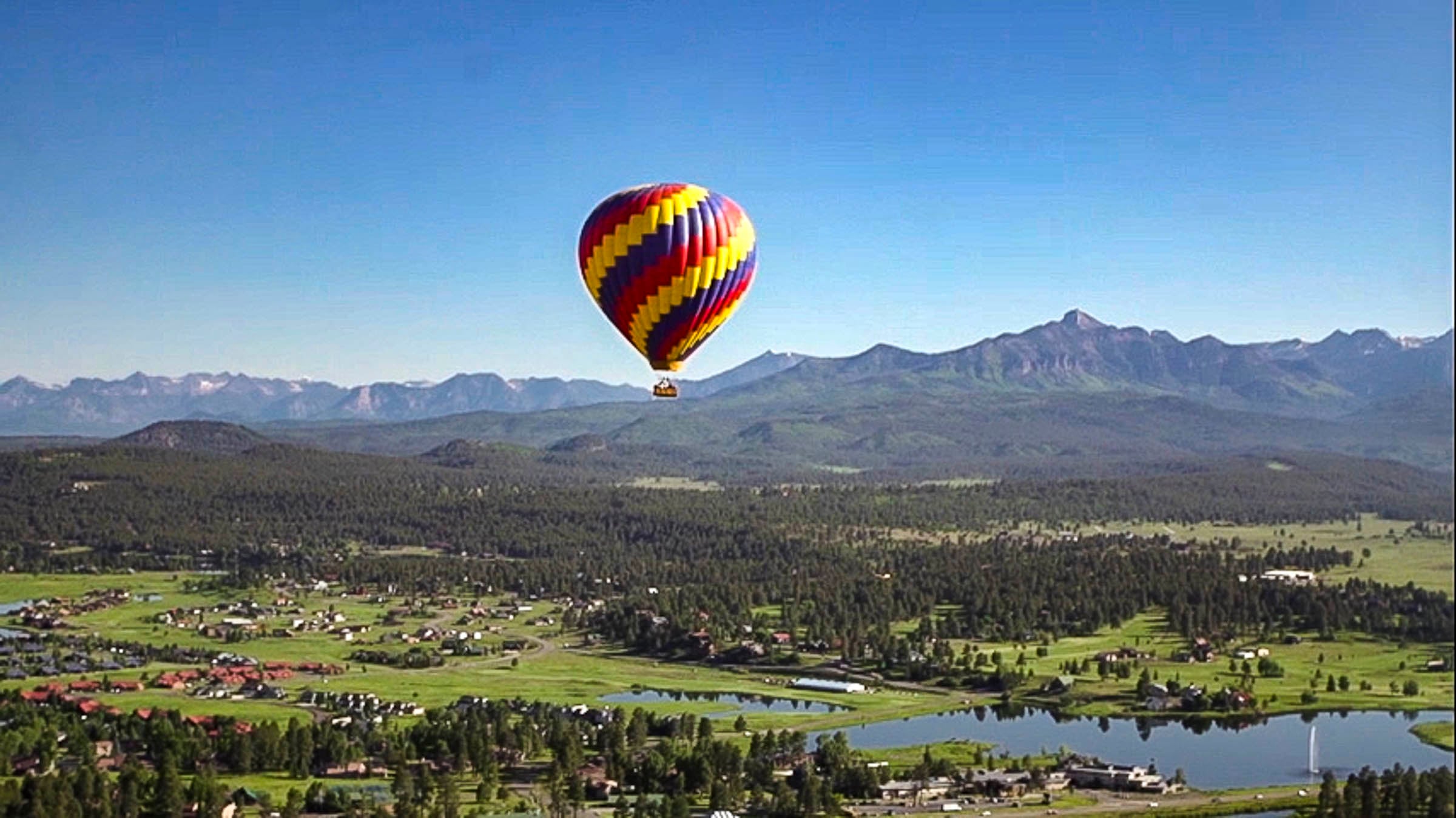 A colorful hot air balloon floats above a mountain valley featuring a town, evergreen forests, and lakes, with distant snow-capped mountains under a clear blue sky.