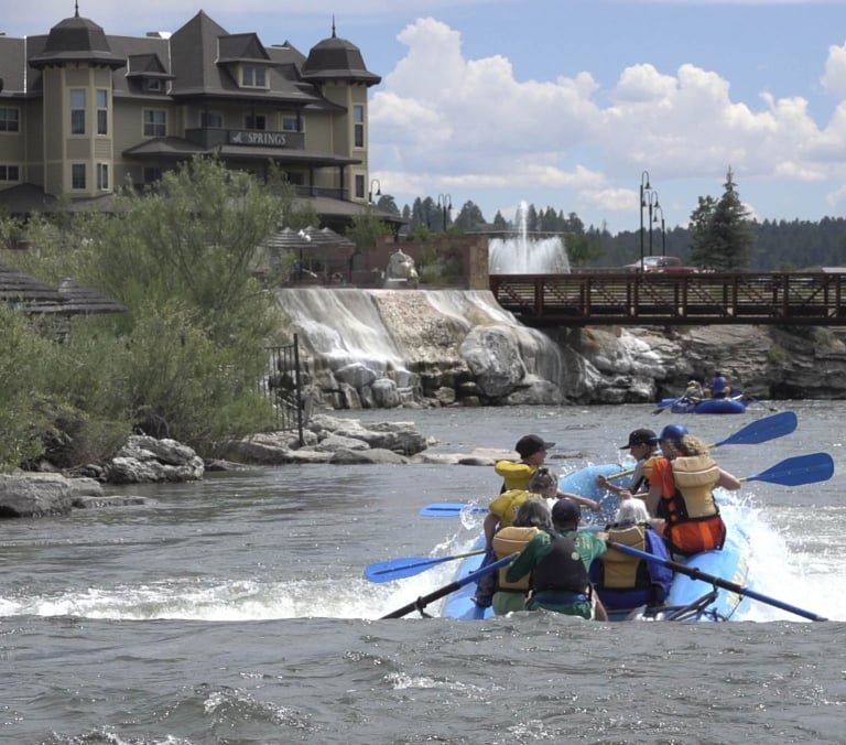 A blue raft with several people paddles through river rapids. Another raft, a resort with **SPRINGS**, a waterfall, and a bridge fill the background.