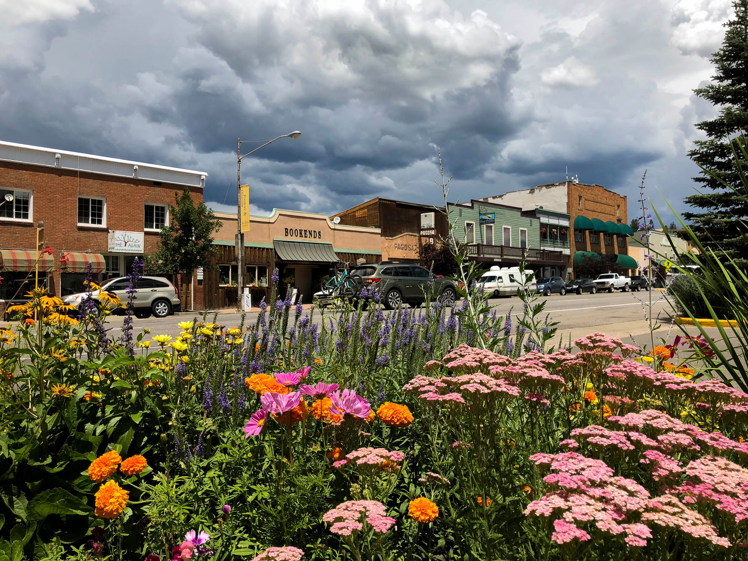 A vibrant flower bed frames a street with parked cars and diverse buildings under a cloudy sky. Shop signs include: HOME AGAIN, BOOKENDS, SUN, ON TAP, PAGOSA, Foot Prints, and a yellow banner reading Welcome & Discover Pagosa.