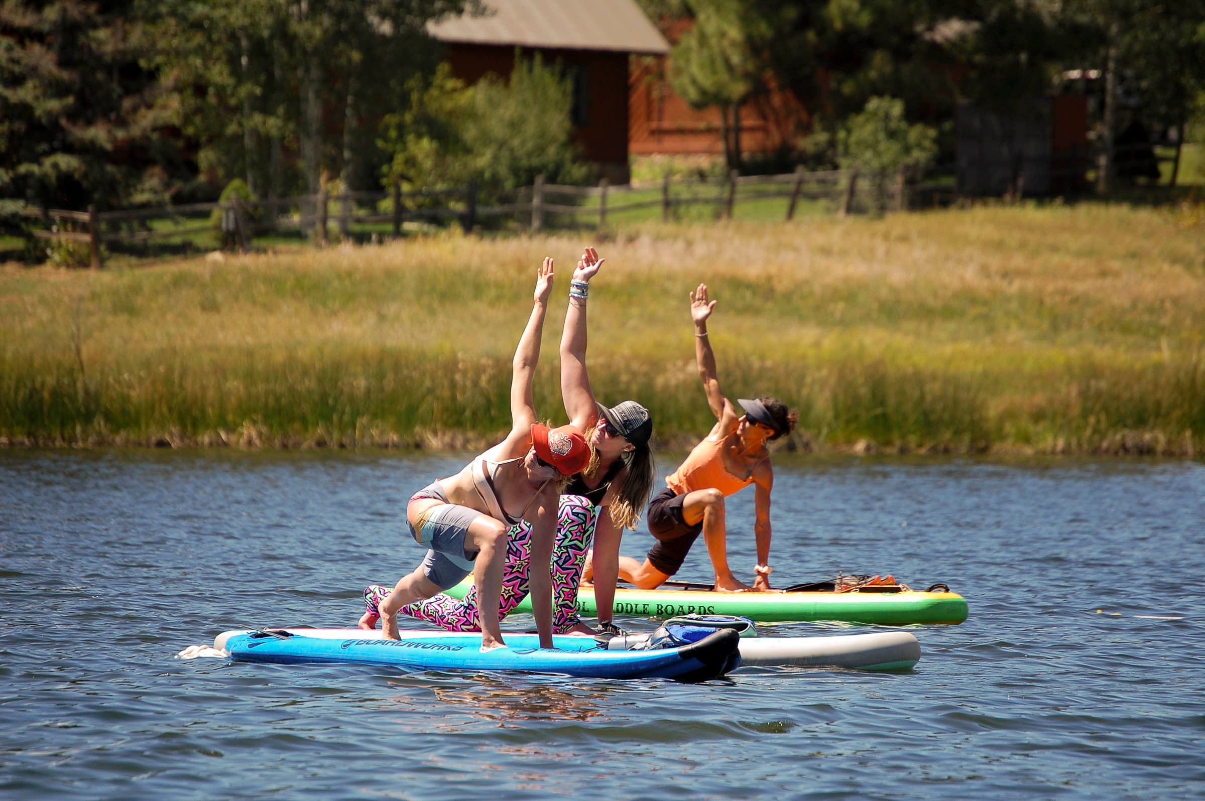 Three women perform yoga lunges with raised arms on paddleboards on a calm lake. Tall grass and a building line the distant shore. Text: 