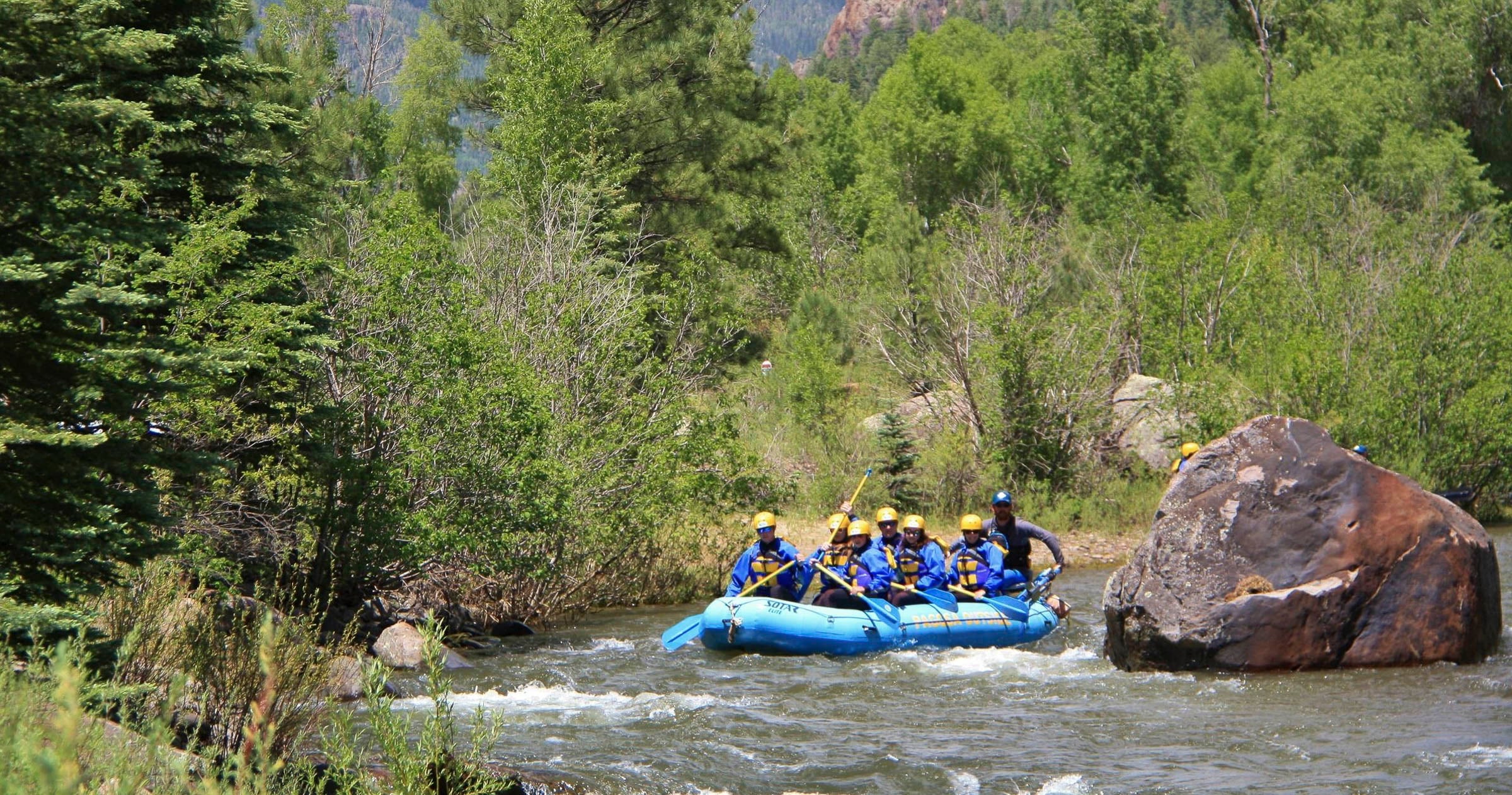 A blue raft carrying six people in yellow helmets and blue vests paddles through rapids, past a large rock and lush green trees along a river. SOTAR ELITE.