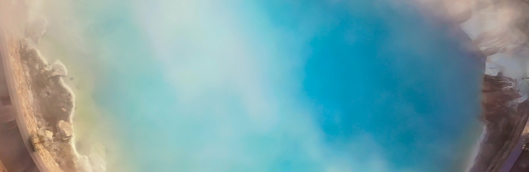 A turquoise-blue geothermal pool steams heavily, with vapor obscuring much of its surface. Rocky, earthy banks and a light-colored path frame the water from an aerial perspective.