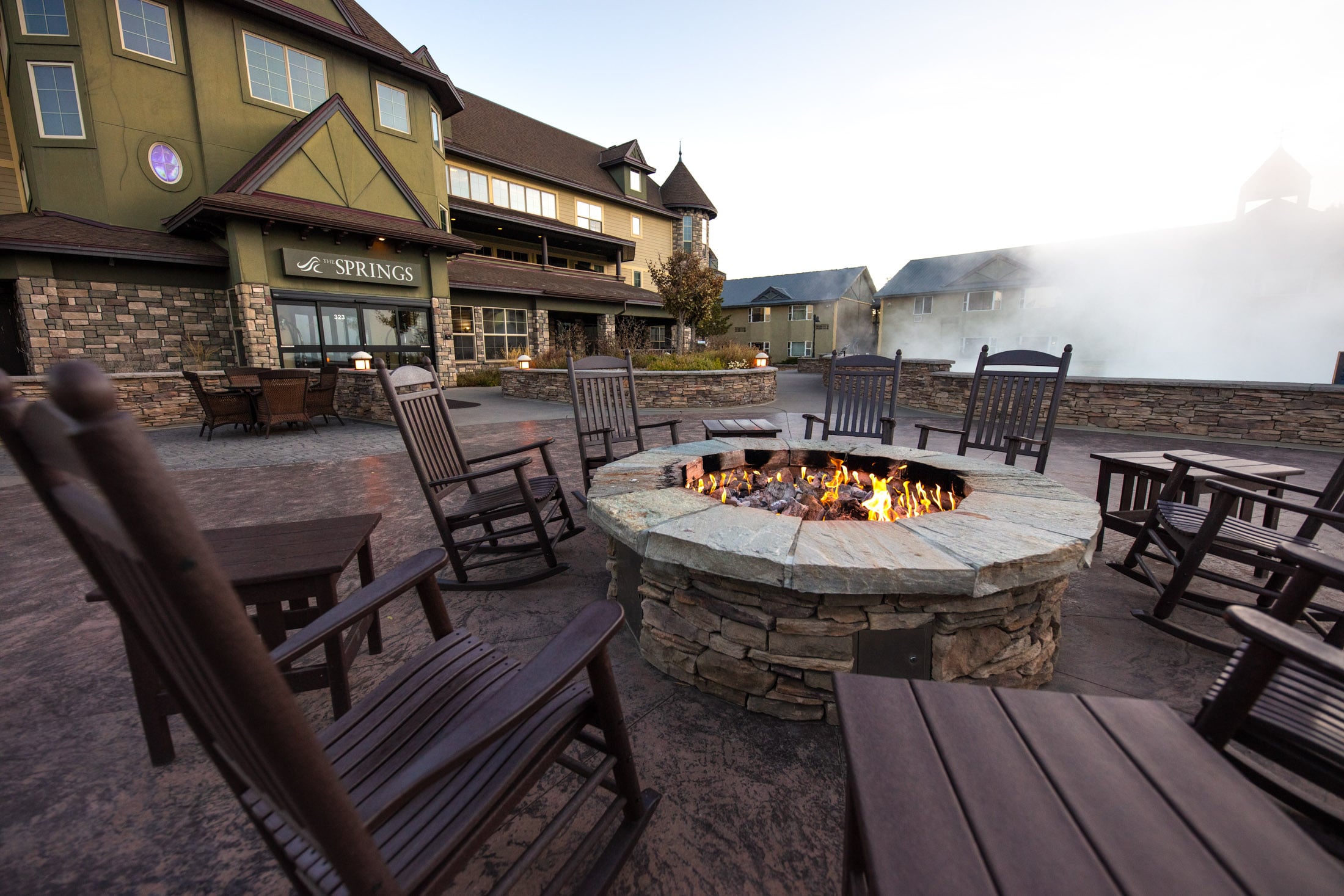 A stone fire pit with burning flames is surrounded by dark rocking chairs on a patio in front of a hotel, 