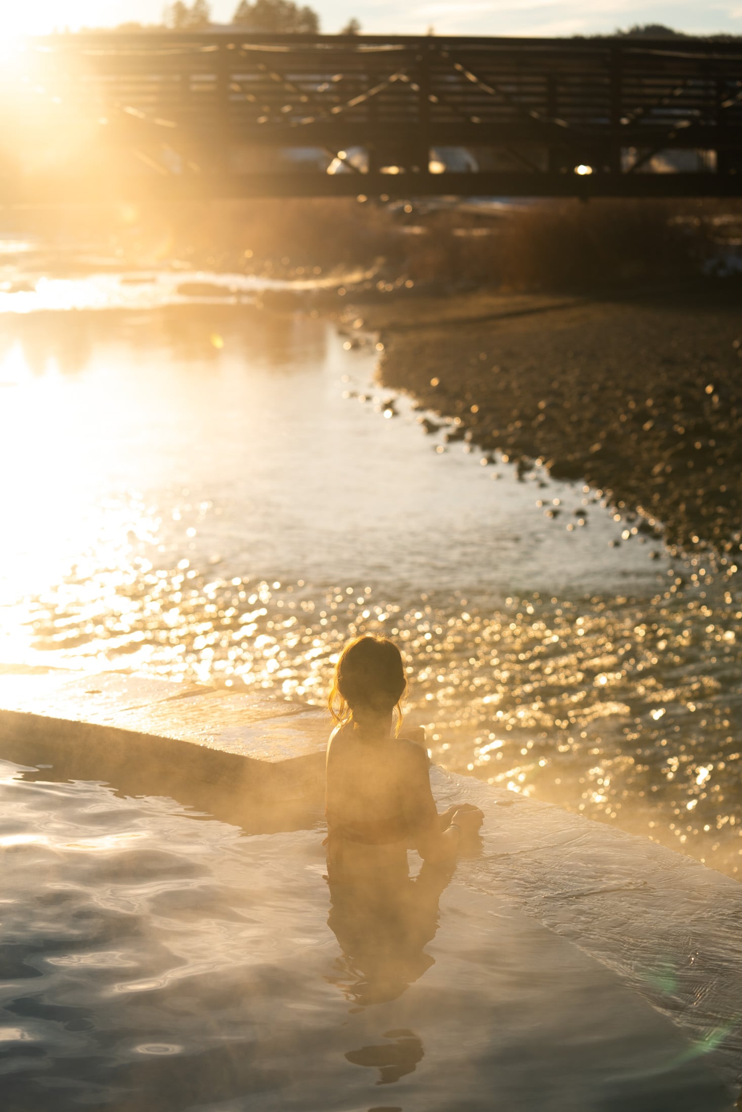 A person sits in a steamy pool, facing a sparkling river with a distant bridge, all illuminated by bright golden hour sunlight.