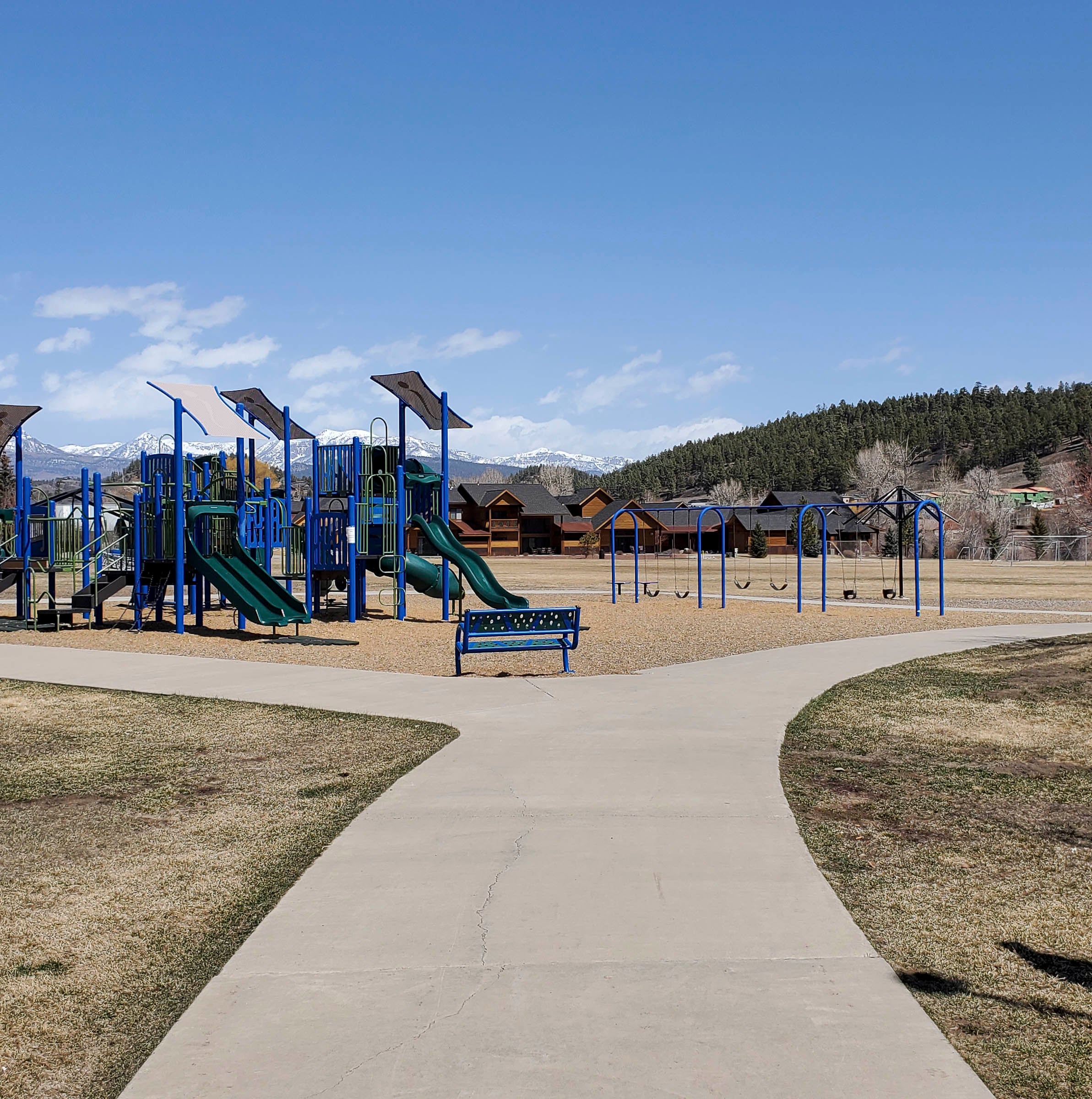 Blue and green playground equipment and swings stand empty in a park. A paved path forks towards distant snow-capped mountains, evergreen forests, and cabins under a clear blue sky.
