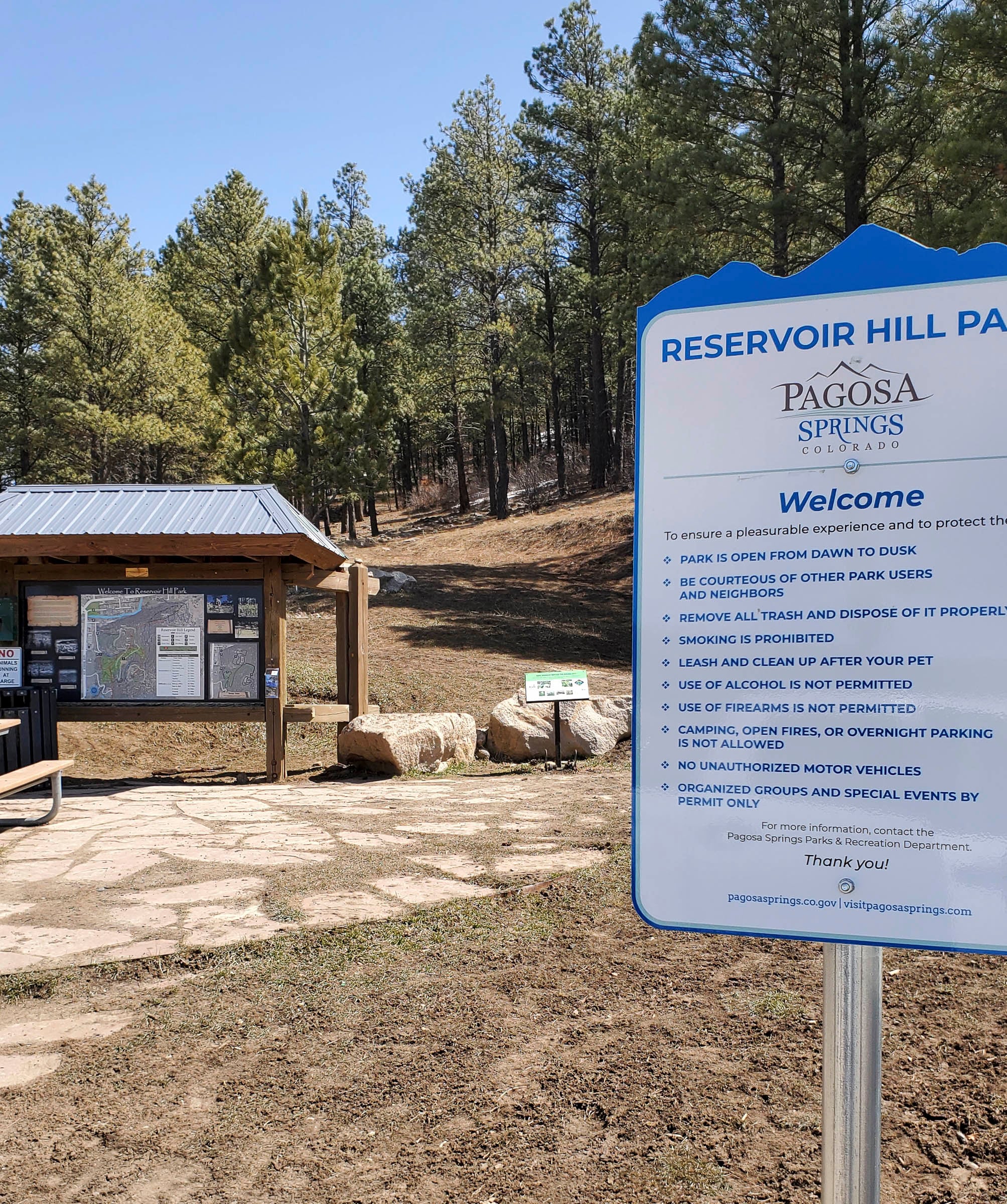 A large park rules sign stands at the entrance to a pine forest, with a wooden information kiosk and flagstone path visible under a blue sky.